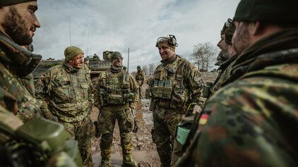 Eine Gruppe männlicher Soldaten der Bundeswehr befindet sich vor einem gepanzerten Fahrzeug im freien Geländer auf matschigem Untergrund und macht eine Lagebesprechung.