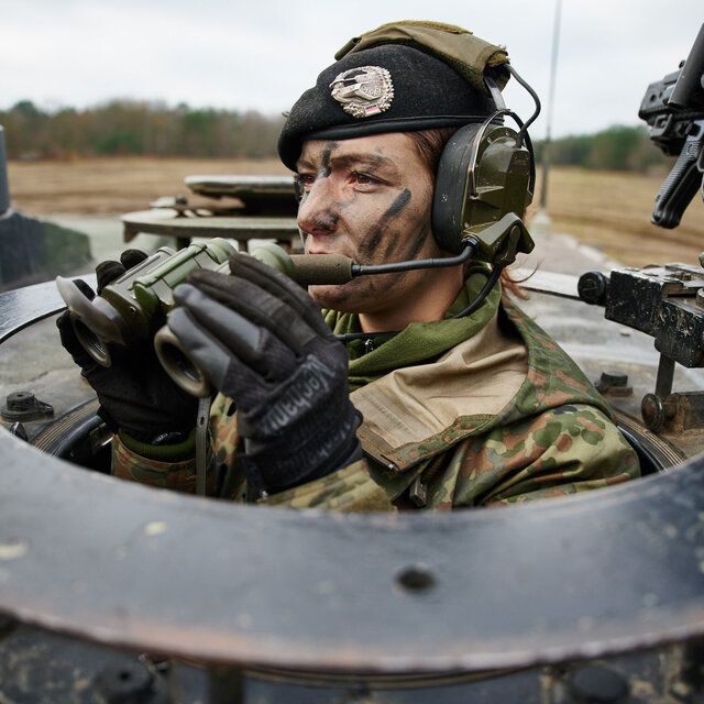 Eine Soldatin beobachtet mit einem Fernglas die Lage aus einem Panzer. Sie trägt Tarnschminke und ihr Barett.