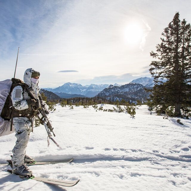 Ein Gebirgsjäger mit Ausrüstung und Skiern steht auf einer verschneiten Fläche. 