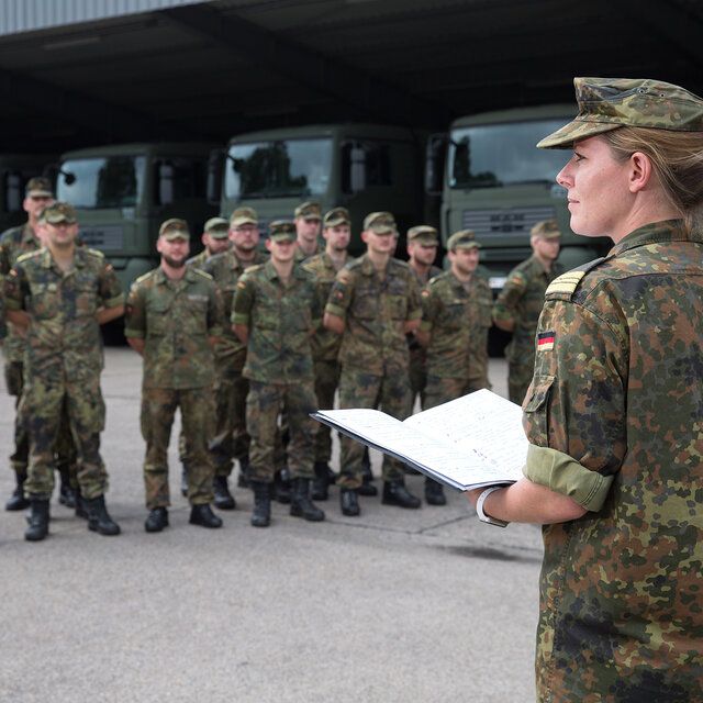 Soldat_innen stehen angetreten vor einer LKW Halle. Eine Offizierin der Marine steht vor der Formation. 